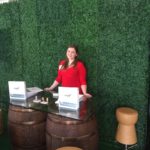 CERF employee promoting Rental Catalog on Wine Barrel Table with Wine Cork Stools and Hedge Backdrop